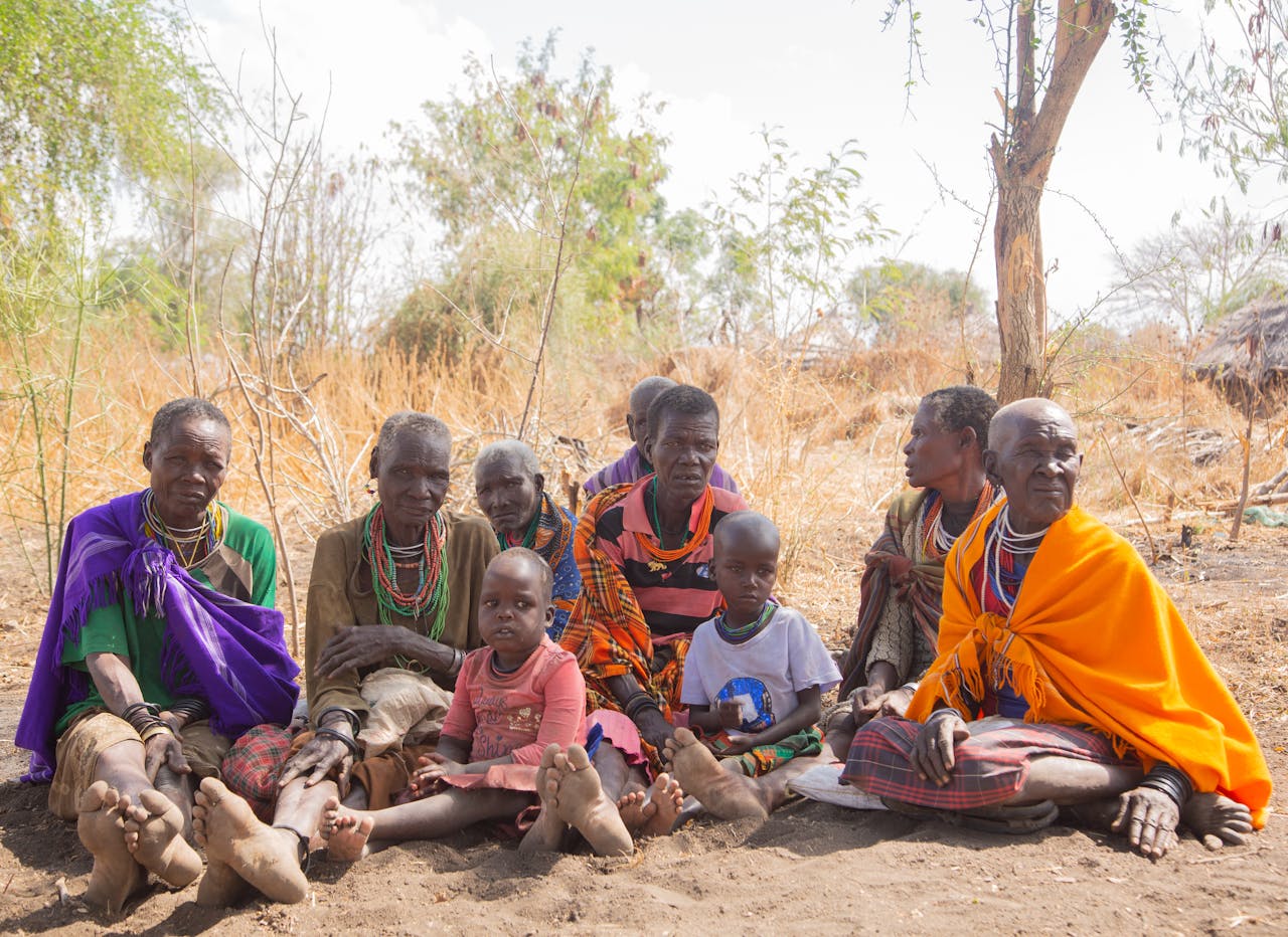 A group of African tribal women and children sitting outdoors in a rural setting.