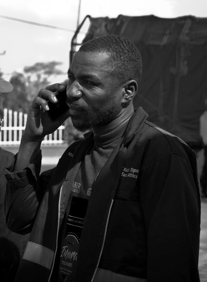 African man talks on phone in Kampala, Uganda, dressed in black jacket, grayscale image.